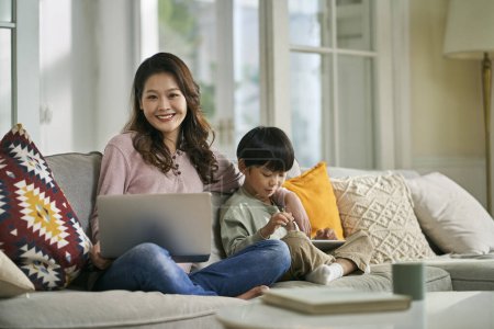 Photo for Beautiful asian mother and five-year-old son sitting on family couch at home looking at camera smiling - Royalty Free Image