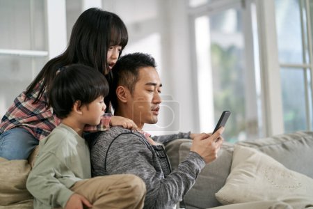 Photo for Young asian father and two children sitting on family couch at home looking at mobile phone together - Royalty Free Image