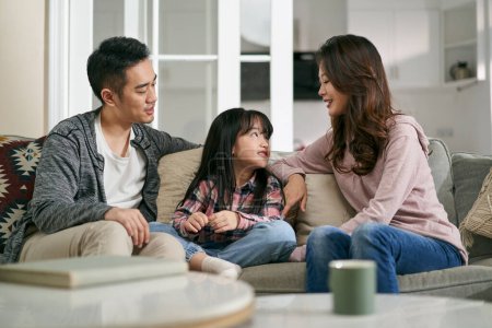 Téléchargez les photos : Jeune asiatique mère et père assis sur la famille canapé à la maison avoir une agréable conversation avec fille - en image libre de droit