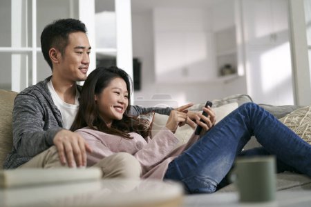 Téléchargez les photos : Heureux jeune asiatique couple assis sur la famille canapé à la maison en regardant des photos de téléphone portable ensemble - en image libre de droit