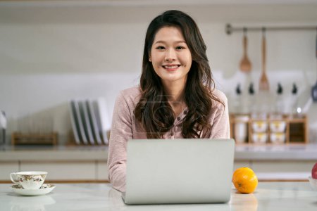 Téléchargez les photos : Portrait d'une femme heureuse et réussie pigiste debout derrière le comptoir de la cuisine à la maison en regardant la caméra sourire - en image libre de droit
