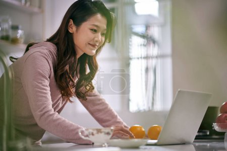 Foto de Joven asiático negocios mujer mujer freelancer trabajando en cocina en casa usando laptop ordenador - Imagen libre de derechos