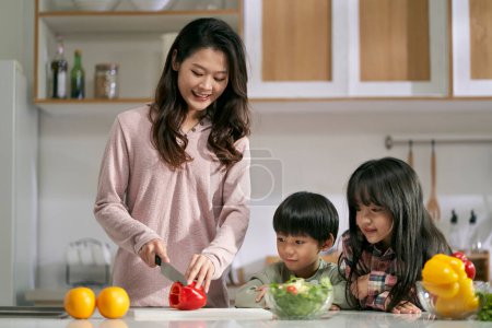Téléchargez les photos : Jeune mère asiatique coupe un poivron pour faire de la salade tandis que ses deux enfants regardent à ses côtés - en image libre de droit