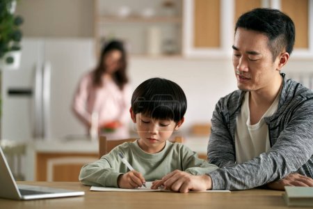 Téléchargez les photos : Jeune asiatique père assis à table tutorat fils à la maison tandis que mère cuisine dans la cuisine - en image libre de droit