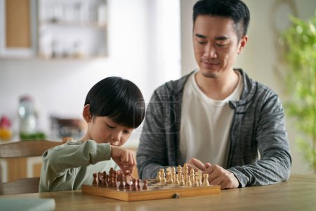Téléchargez les photos : Asiatique père et fils assis à cuisine table à la maison jouer aux échecs - en image libre de droit