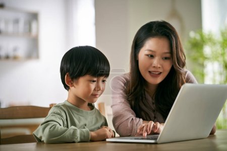 Photo for Happy asian mother and son sitting at table at home watching movie using laptop computer - Royalty Free Image