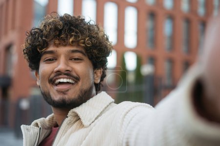 Lächelnder moderner indischer Mann, der ein Selfie macht, Videoanrufe auf der Straße macht. Emotionale Lockenkopf asiatischen Influencer Aufnahme von Video, Kommunikation im Freien. Funktechnologiekonzept 