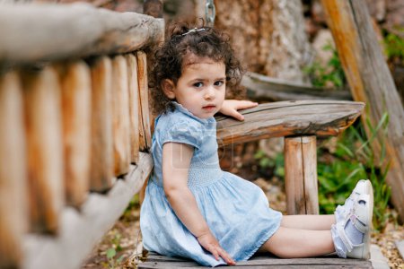 Curly-haired two-year-old girl sitting on a wooden bench in the park and looking at camera