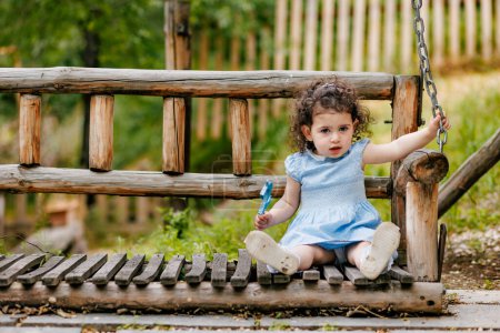 Outdoor photoshoot. Cheerful child in blue dress on a wooden swing in a green park and holding her birthday number candle