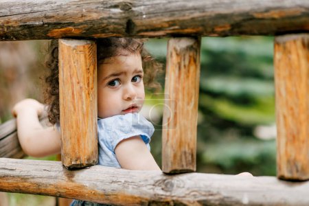 Nachdenklich lockiges Kleinkind sitzt auf einer handgefertigten Holzbank und blickt traurig in die Kamera