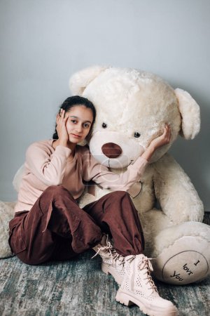 Linda chica de moda de 13 años sentada en el suelo con su peluche y posando para la cámara en el estudio