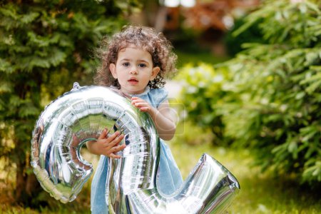 Birthday photoshoot. Sweet little girl with curly hair holding balloon number in the garden, looking at camera carefully