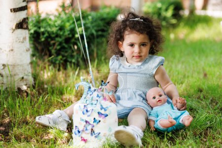 Young child with curly hair sitting on grass beside birch tree with her doll and birthday cake