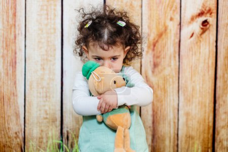 A tender scene. A pensive little girl cuddles her stuffed teddy in front of wooden background.