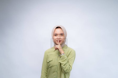 Do not be noisy. Portrait of young asian woman wearing hijab keeps his finger in his mouth at Copy Space Advertising Your Text, Standing Isolated Over white Studio Background