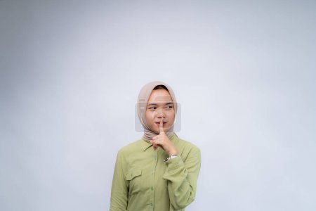 Do not be noisy. Portrait of young asian woman wearing hijab keeps his finger in his mouth at Copy Space Advertising Your Text, Standing Isolated Over white Studio Background