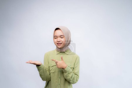 Smiling beautiful Asian woman wearing hijab pointing finger sideward to empty space in studio white isolated background