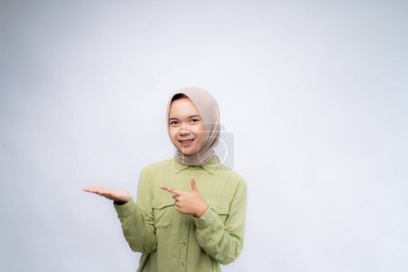 Smiling beautiful Asian woman wearing hijab pointing finger sideward to empty space in studio white isolated background