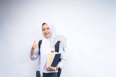 Eine indonesische Gymnasiastin in weiß-grauer Uniform, die eine Tasche trägt und ein Buch in der Hand hält, lächelt und zeigt OK-Zeichen