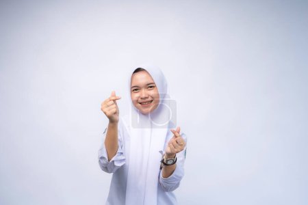 Eine indonesische High-School-Schülerin in weiß-grauer Uniform, die mit dem Finger ein Liebeszeichen zeigt, steht isoliert über weißem Studiohintergrund.