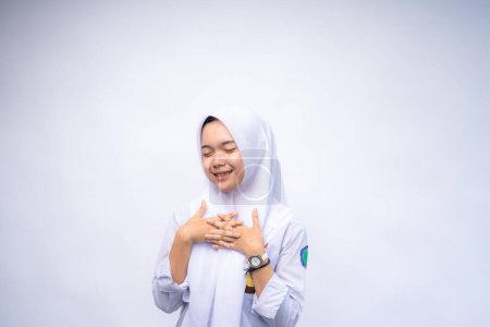 Glücklich achtsame, dankbare indonesische Gymnasiastin in weiß-grauer Uniform, die lächelnd die Hände auf der Brust hält. Bildungskonzept