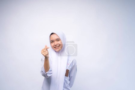 Téléchargez les photos : Femme indonésienne lycéenne en uniforme blanc et gris montrant l'amour coréen signe avec le doigt à l'espace de copie annonçant votre texte, debout isolé sur fond de studio blanc - en image libre de droit