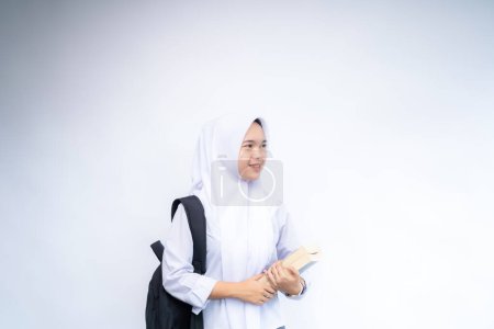 Weibliche indonesische High-School-Studentin in weiß-grauer Uniform hält ein Buch an der Copy Space Advertising Your Text, steht isoliert über weißem Studiohintergrund