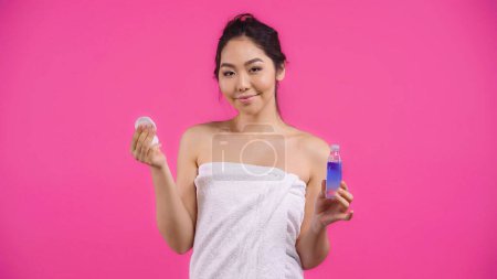 asiatische junge Frau in Handtuch hält Baumwollkissen und micellar Wasser isoliert auf rosa 