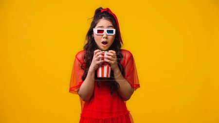 shocked asian woman in 3d glasses holding popcorn and watching scary movie isolated on yellow 