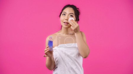 Téléchargez les photos : Asiatique jeune femme dans serviette tenant coton pad avec micellaire eau tout nettoyage visage isolé sur rose - en image libre de droit