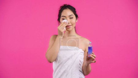 junge asiatische Frau in Handtuch hält Baumwollkissen mit Mikrokellerwasser, während Reinigung Gesicht isoliert auf rosa 