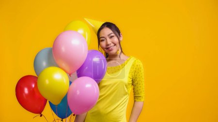 happy asian woman in party cap holding colorful balloons isolated on yellow 