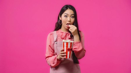Foto de Joven asiático mujer con abierto boca comer salado palomitas cubo aislado en rosa - Imagen libre de derechos