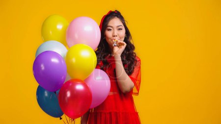 asiático mujer en rojo vestido celebración colorido globos y soplado parte cuerno aislado en amarillo 