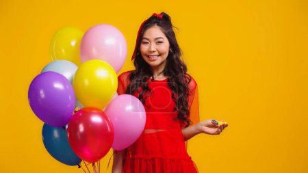cheerful asian woman in red dress holding colorful balloons and party horn isolated on yellow 