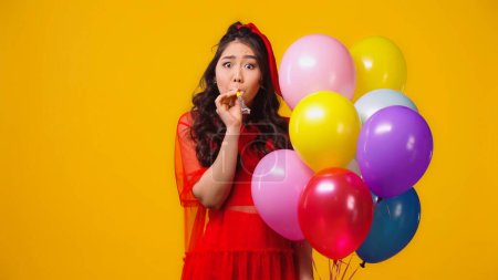 asian woman in red dress holding balloons and blowing party horn isolated on yellow 