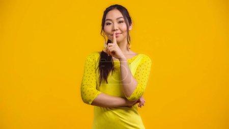 Téléchargez les photos : Heureux asiatique femme montrant silence signe et sourire isolé sur jaune - en image libre de droit
