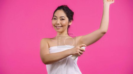 asian and happy woman in towel applying deodorant isolated on pink 