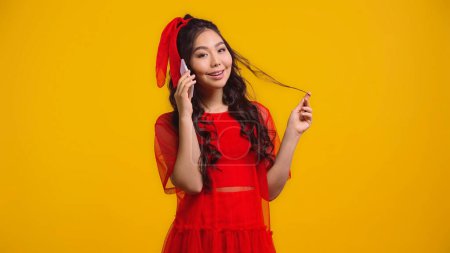 sonriente asiático mujer en rojo vestido hablando en smartphone mientras girando pelo aislado en amarillo 