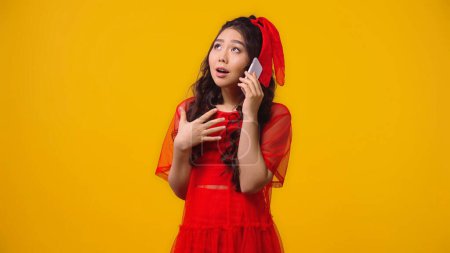 Téléchargez les photos : Femme asiatique émotionnelle en robe rouge parler sur smartphone isolé sur jaune - en image libre de droit