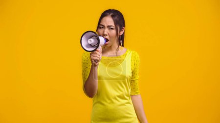 displeased asian woman screaming in megaphone isolated on yellow 
