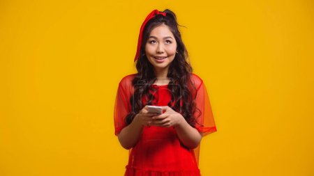 Téléchargez les photos : Heureux asiatique femme en rouge robe textos sur smartphone isolé sur jaune - en image libre de droit