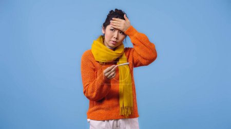 kranke asiatische Frau in Pullover und Schal mit digitalem Thermometer und berührender Stirn isoliert auf blauem Grund 