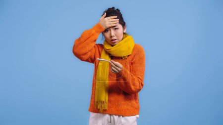 kranke asiatische Frau in Pullover und Schal hält digitales Thermometer und berührt Stirn isoliert auf blauem Grund 