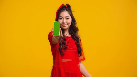 cheerful asian woman holding smartphone with green screen isolated on yellow 