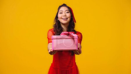 positive and young asian woman giving wrapped present isolated on yellow 