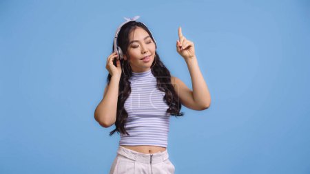 Téléchargez les photos : Heureux asiatique femme dans sans fil casque écoute musique et pointage avec doigt isolé sur bleu - en image libre de droit