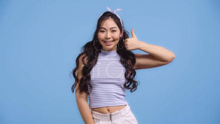 happy asian woman in hoop earrings showing thumb up isolated on blue 