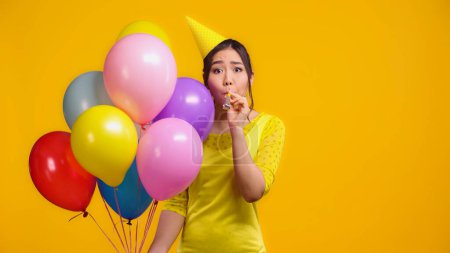 asian woman holding colorful balloons and blowing party horn isolated on yellow 