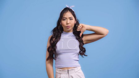 dissatisfied asian woman in crop top showing thumb down isolated on blue  